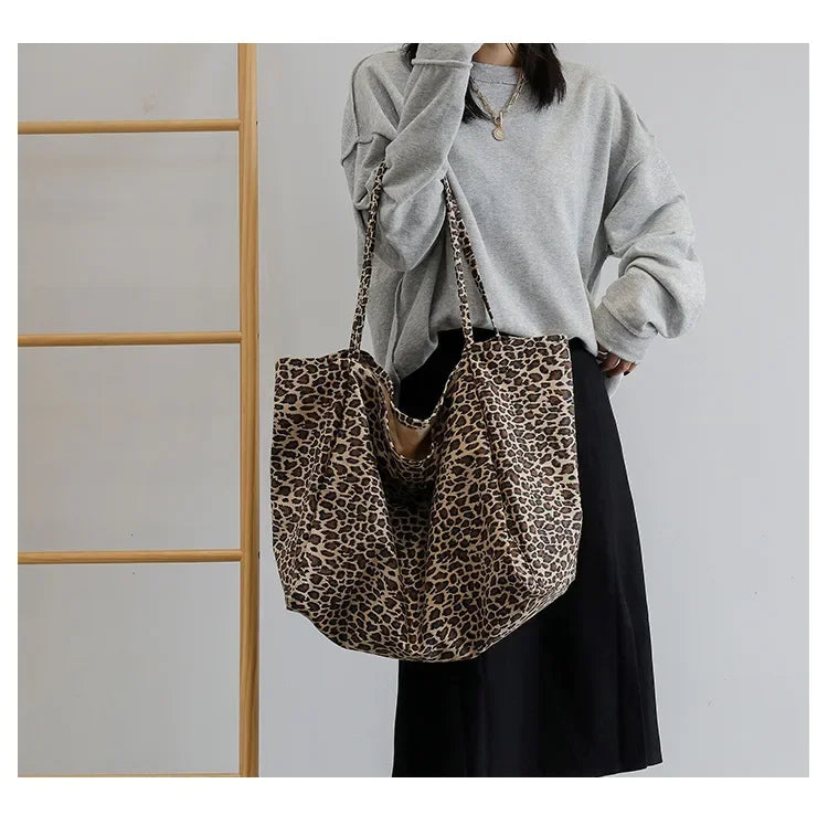A woman holds a leopard print canvas crossbody tote bag with brown leather straps against a light gray sweater and black skir