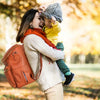 A woman is laughing while holding a child on her shoulder outdoors with an orange backpack.