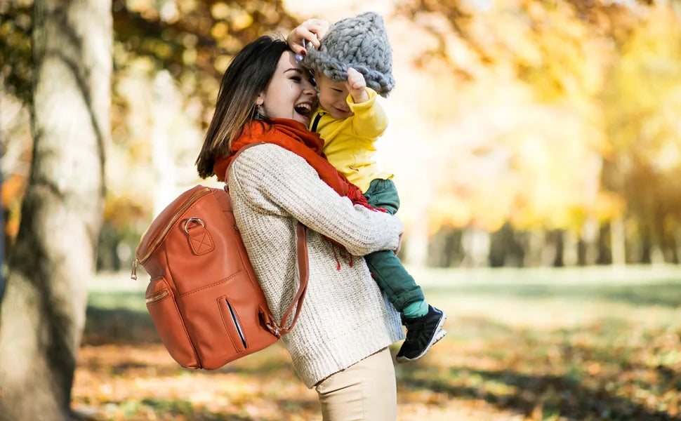 A woman is laughing while holding a child on her shoulder outdoors with an orange backpack.