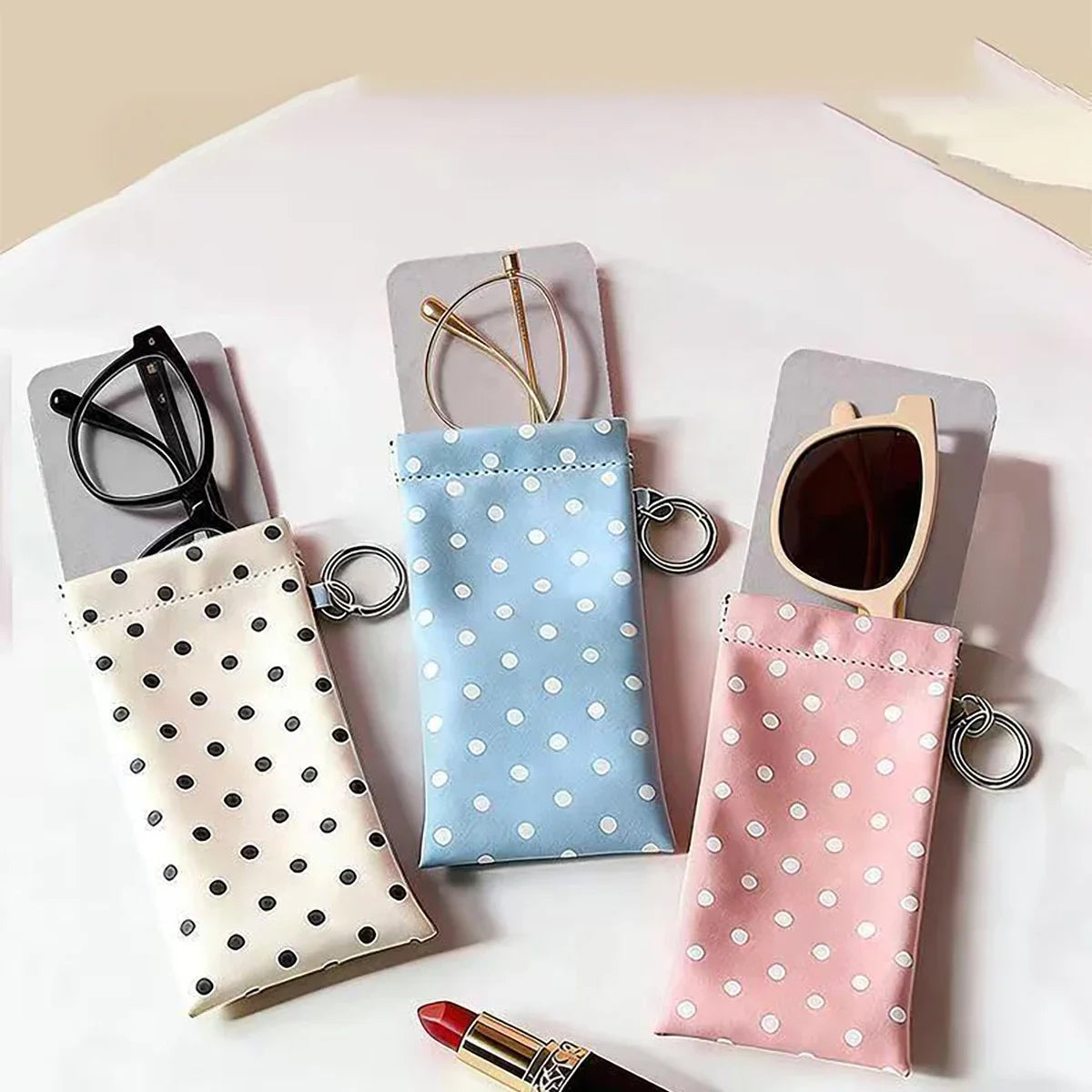 A set of pink, blue, and beige polka dot eyeglass pouches with metal keychains displayed on a white surface.