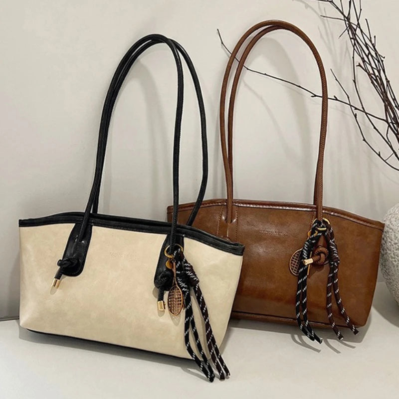 A beige leather tote bag with black leather handles and strap details is displayed next to a brown leather tote bag.
