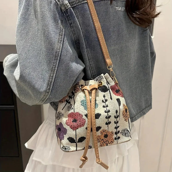 A close-up of a floral drawstring bucket bag with light beige fabric and leather accents worn on a person's shoulder.