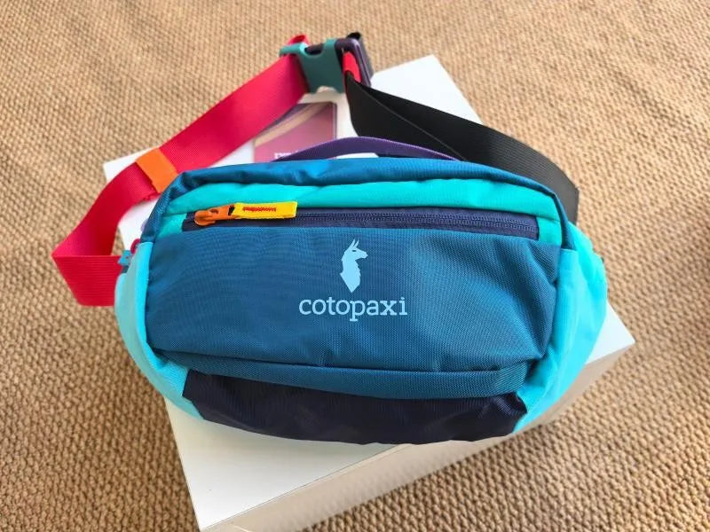 A teal and navy patchwork color block fanny pack with a black strap resting on a white box.