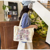 A woman wearing a denim shirt and apron holds a transparent shoulder tote bag in a bright indoor setting.