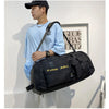 A young man wearing a light blue shirt holds a black tactical backpack against a white wall.