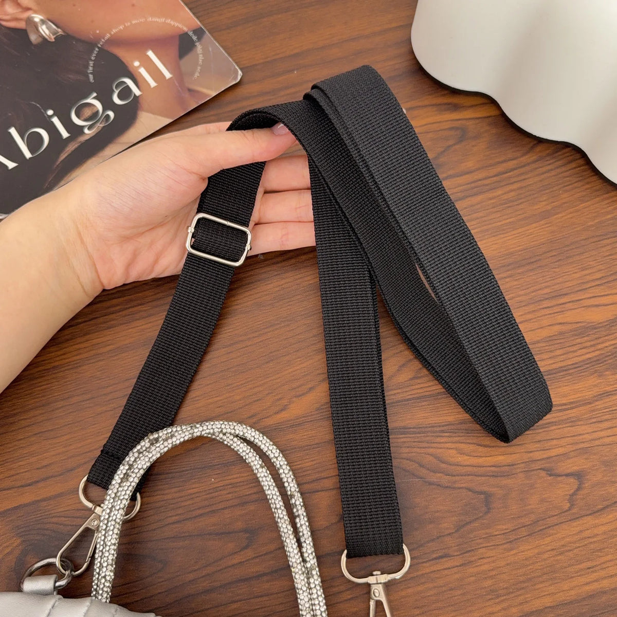 A black adjustable wide shoulder strap is shown next to a silver chain strap on a wooden surface.