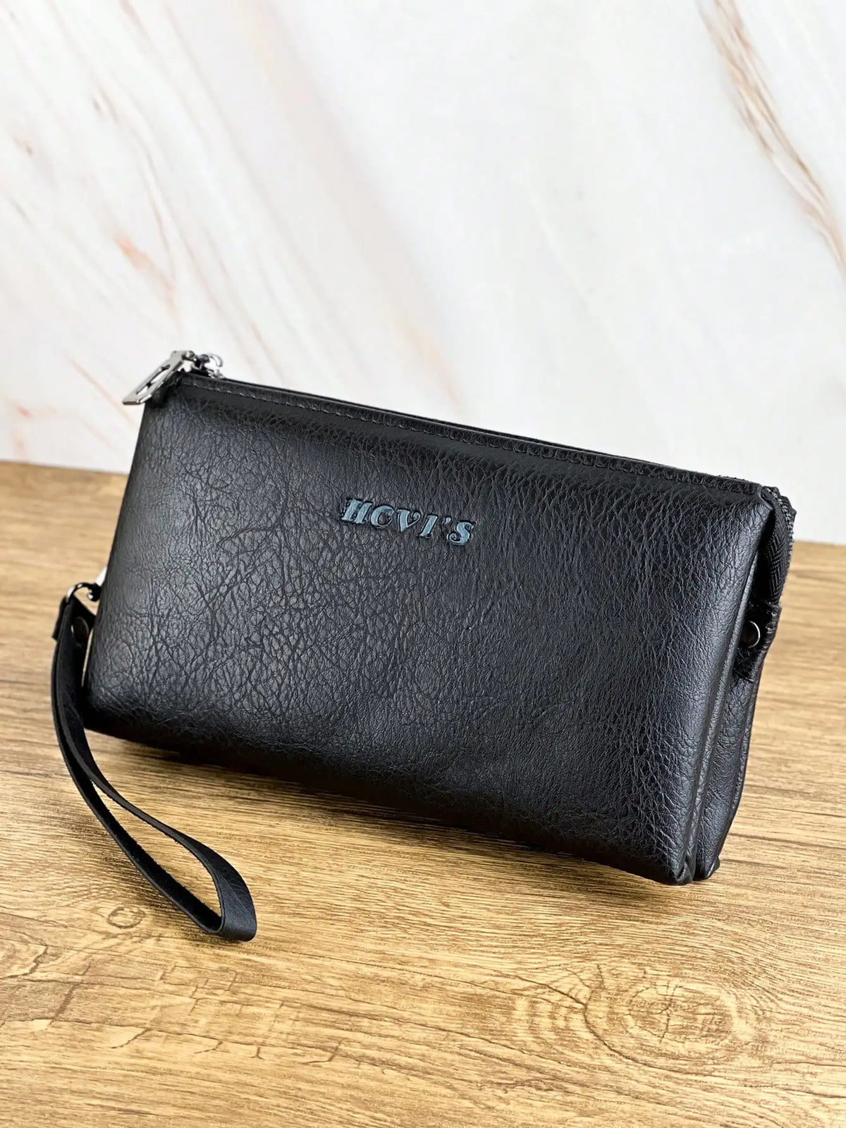 Portefeuille pochette en cuir noir avec quincaillerie argentée posé sur une surface en bois.