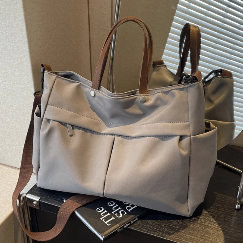 A beige lightweight nylon tote bag with brown leather handles and multiple exterior pockets resting on a dark surface.