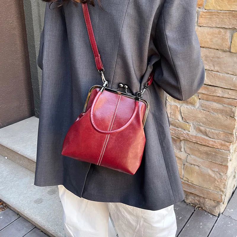 A woman is holding a red leather shell handbag with a strap against a brick wall.