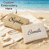 Two handwoven straw makeup bags, one labeled Sophia and the other Camille, are placed on sandy beach with seashells.