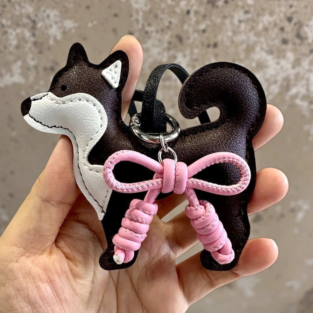 Un dije de perro Shiba Inu negro con marcas blancas y un lazo de cuero trenzado rosa sostenido en una mano.