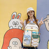 A young woman wearing a white cap and a beige backpack stands in front of colorful wall art featuring cartoon rabbits and fig
