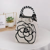 A beige knitted bucket bag with a black and white floral design and black patterned handles sits on a white table.