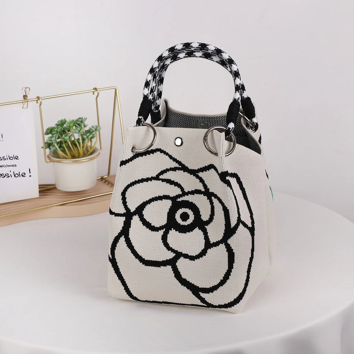A beige knitted bucket bag with a black and white floral design and black patterned handles sits on a white table.