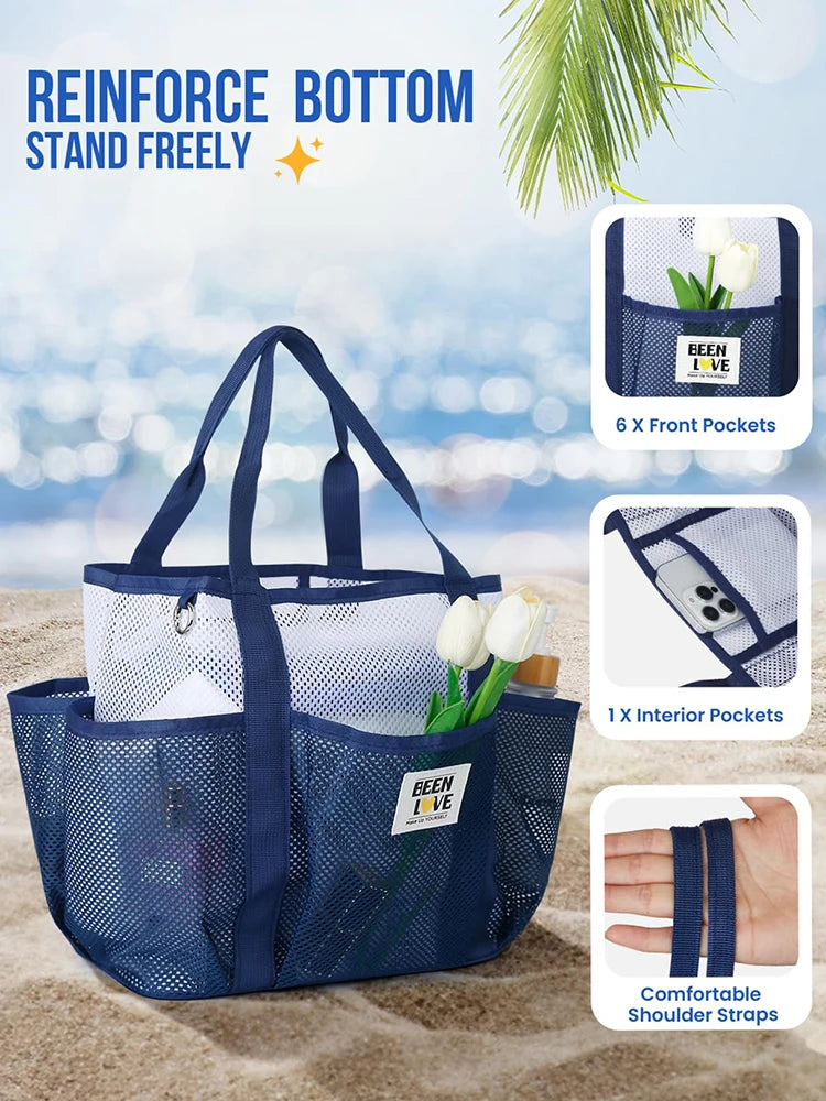 A navy blue mesh beach tote bag with shoulder straps and front and interior pockets resting on sand.