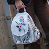 A light gray linen backpack featuring floral embroidery in pink and blue tones, carried by a person.