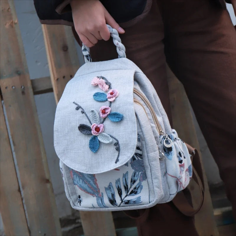 A light gray linen backpack featuring floral embroidery in pink and blue tones, carried by a person.