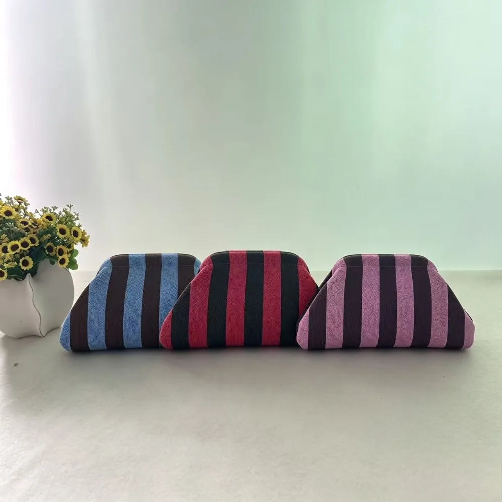 A row of three striped fabric makeup bags in blue, maroon, and purple are displayed on a white surface next to a potted plant