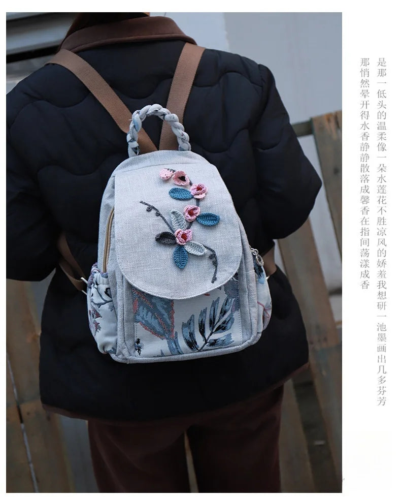 A woman wearing a dark coat carries a light gray linen backpack with pink and blue 3D embroidery.