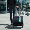 A person in a black suit is pulling a black rolling suitcase with a luggage tag across a paved outdoor area.
