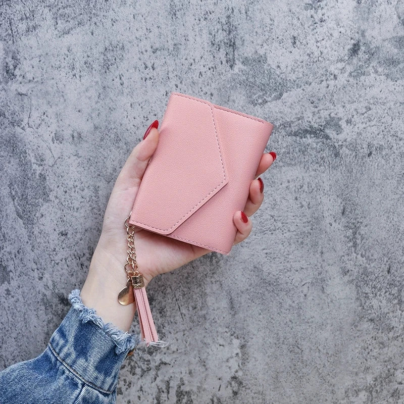 A hand holds a pink PU leather coin clutch wallet with a gold tassel against a gray textured background.