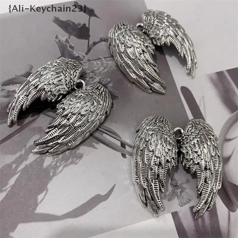 Silver metal angel wing keychain charms resting on a dark background with foliage.