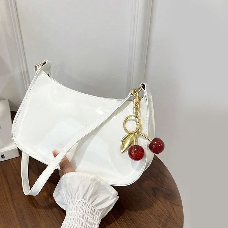 A white PU leather shoulder bag with a gold chain and cherry pendant resting on a wooden surface.