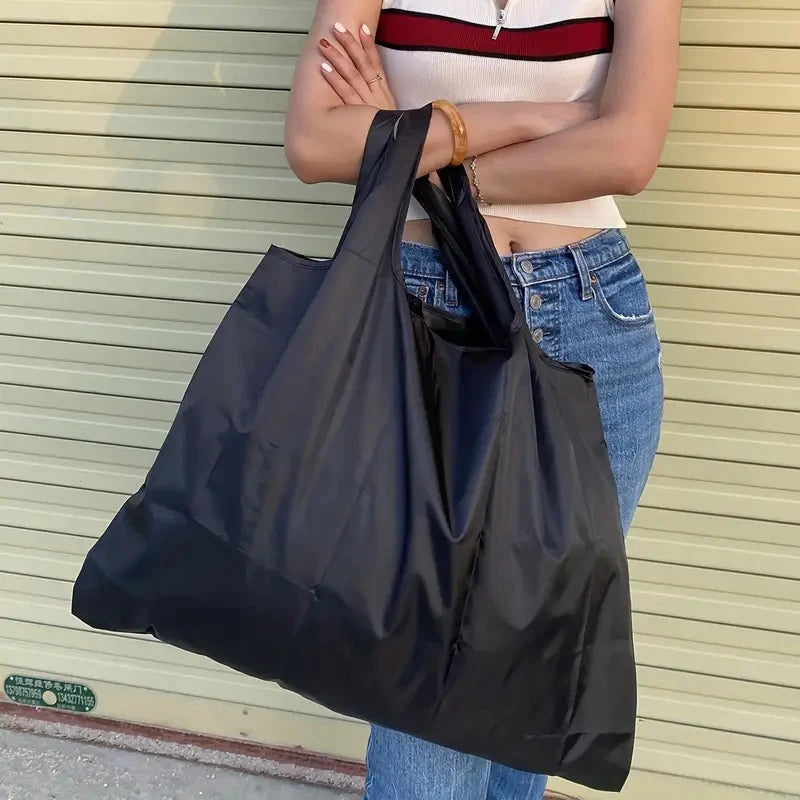 A person holds a black foldable water-resistant shopping tote bag against a light beige corrugated wall.