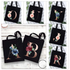 A set of black canvas tote bags featuring an alphabet initial and floral design in rose and blue tones.