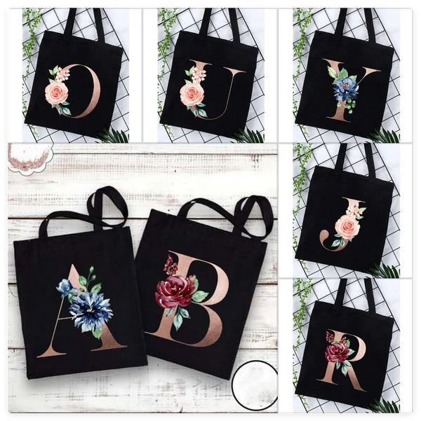 A set of black canvas tote bags featuring an alphabet initial and floral design in rose and blue tones.