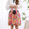 A woman holds a large, colorful floral reusable shopping bag against a white shirt.