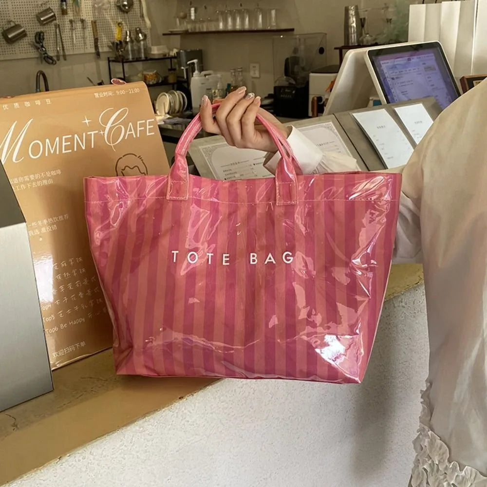 A pink and white striped transparent PVC tote bag is held by a person in a cafe setting.
