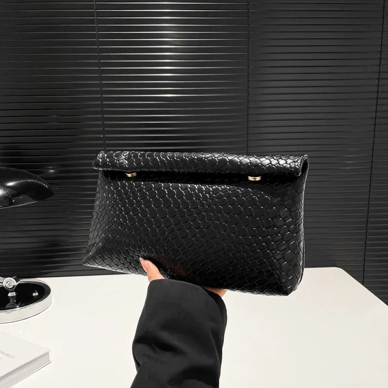 Una mano sostiene un elegante bolso de mano tejido negro contra un fondo oscuro texturizado.
