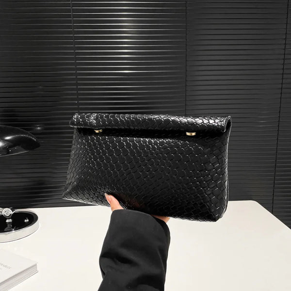 A hand holds an elegant black woven clutch bag against a dark textured background.