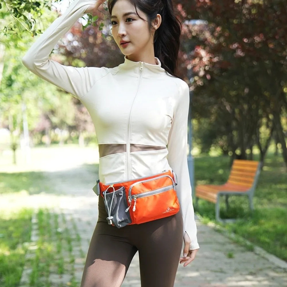A woman wearing a white long-sleeved top and brown pants holds an orange waterproof invisible waist pack outdoors.