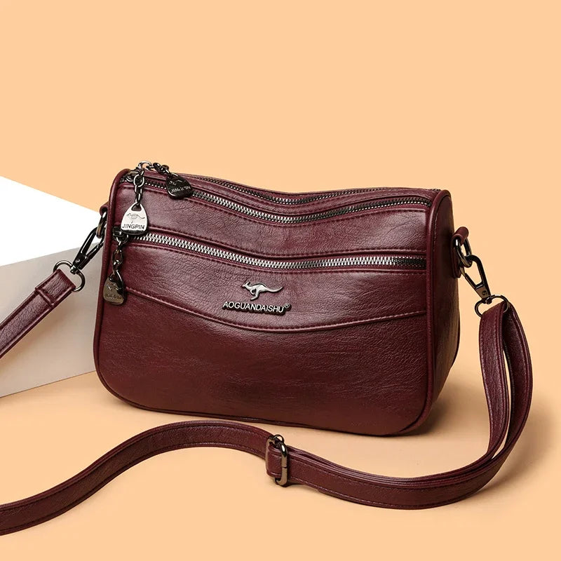 Bolso cruz de cuero marrón borgoña y moderno para mujer con cremallera y correa de cuero sobre fondo claro.