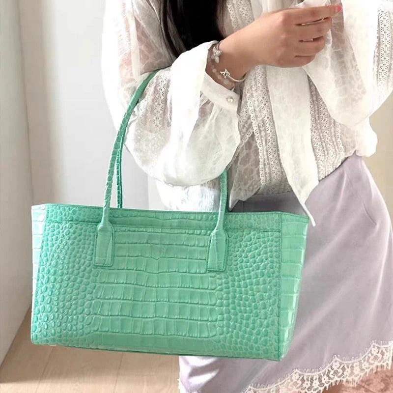 A mint green crocodile print leather handbag carried by a person wearing a white lace top and light purple skirt.