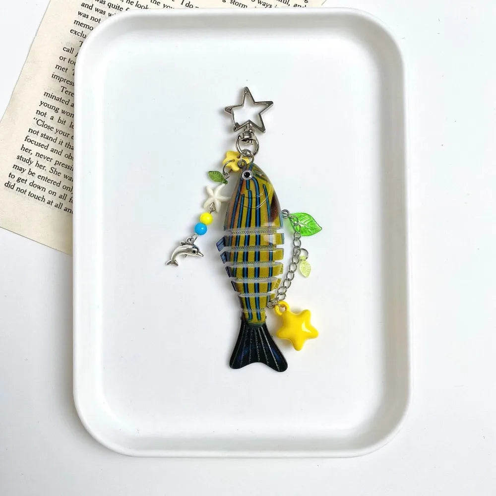 A striped simulation fish keychain with a star charm and yellow accents displayed on a white square dish.