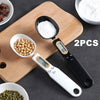 A precision digital kitchen scale is shown with two spoons, measuring white powder and whole peppercorns on a wooden cutting 
