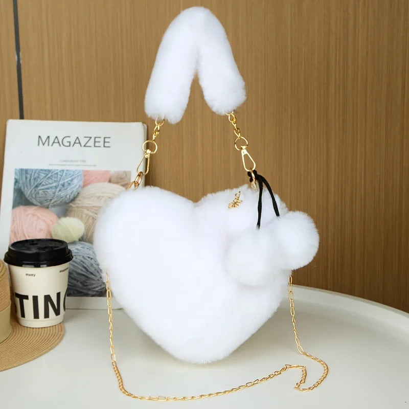 A white faux fur heart shaped crossbody purse with gold chain and black strap resting on a light surface.