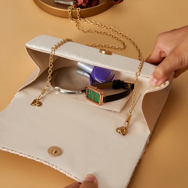A hand holds a satin evening clutch with a gold chain strap and a purple accessory.