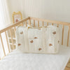 A cream quilted diaper bag tote with small brown bear patches resting on a white surface next to a wooden crib.
