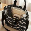 A black tote bag with a white bat print and a black strap, holding a pen and a small notebook.