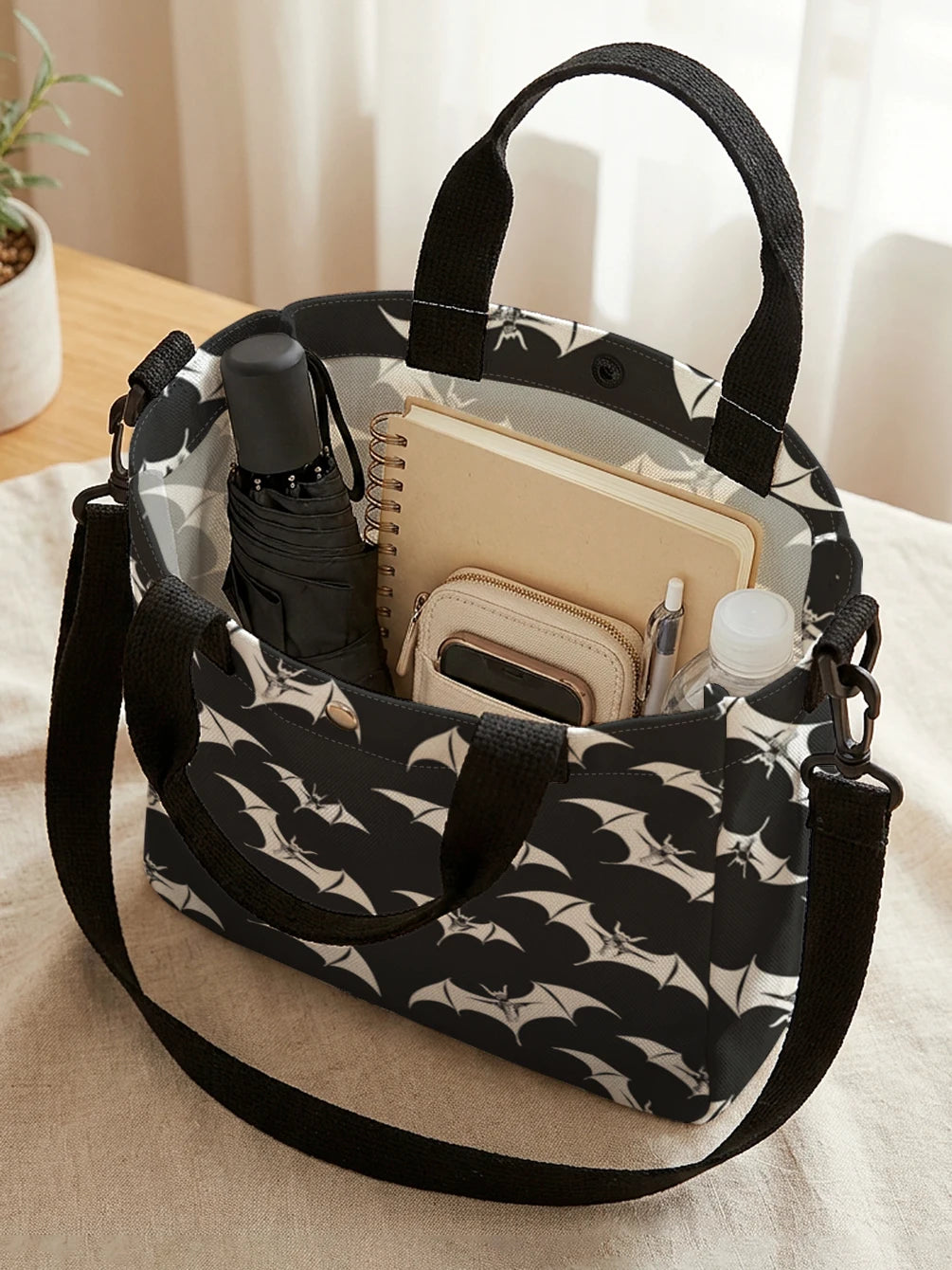 A black tote bag with a white bat print and a black strap, holding a pen and a small notebook.