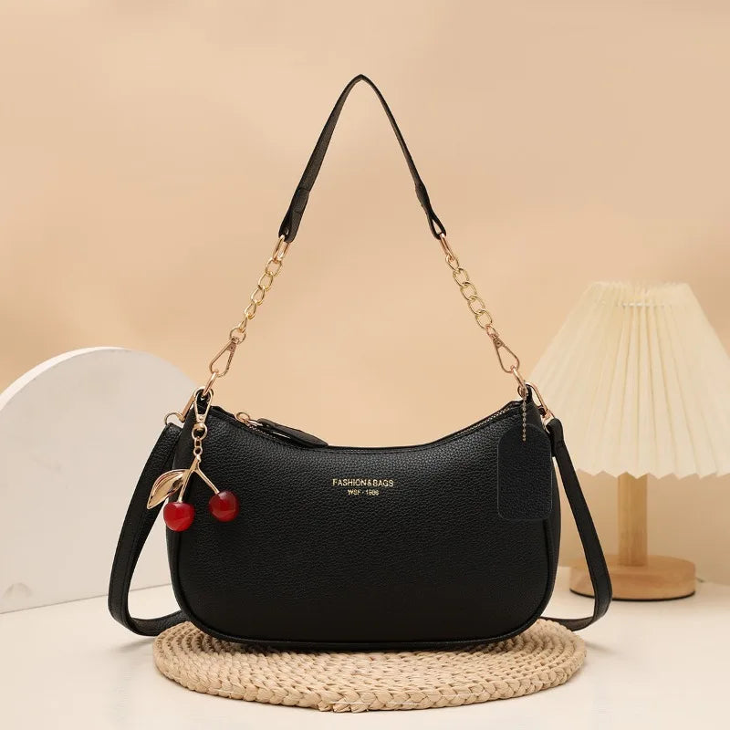 Black leather shoulder bag with a gold chain strap and a red cherry pendant resting on a woven mat.