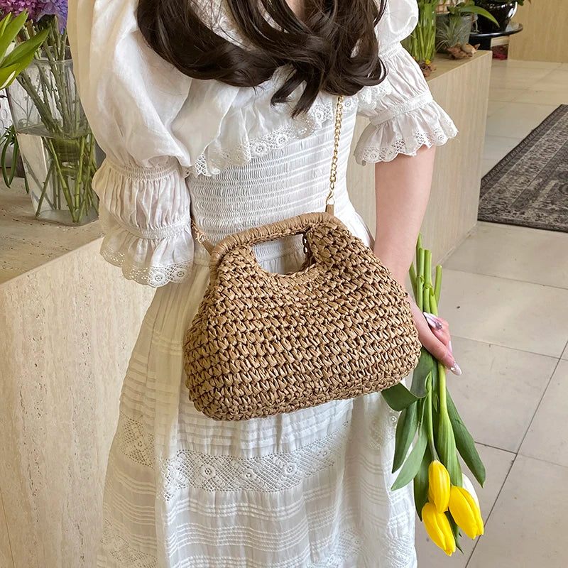 A woman wearing a white dress holds a chain woven straw shoulder bag and a bunch of yellow flowers.