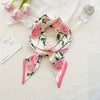 Foulard floral de soie rose et vert noué en nœud, posé sur un tissu blanc à côté d'une bouteille en verre.