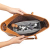 A hand is opening a tan leather handbag to reveal a gray organizer insert containing various small metal and wooden accessori