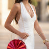 A woman wearing a white dress holds a gold acrylic chain crossbody bag with a red fan in an outdoor setting.