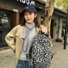 A woman wearing a beige coat and jeans holds a black leopard print canvas backpack outdoors.
